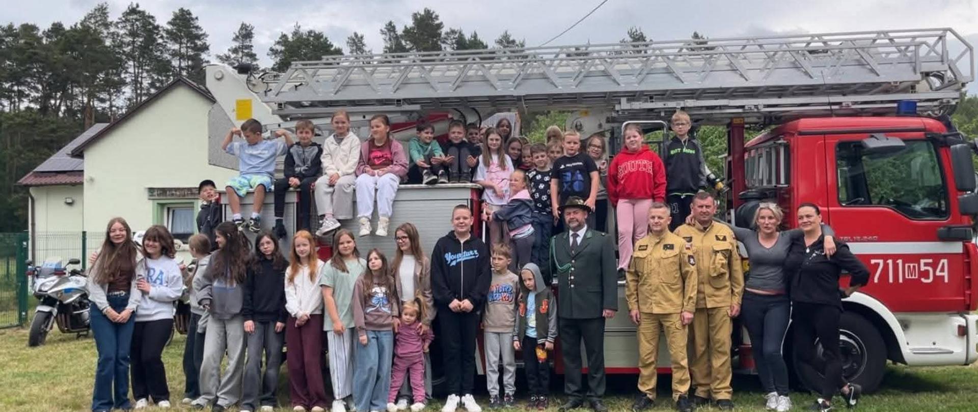 Duża grupa dzieci i kilku dorosłych pozuje do pamiątkowego zdjęcia na tle czerwonego wozu strażackiego z drabiną mechaniczną. Część osób stoi na trawie przed pojazdem, a liczna grupa dzieci siedzi na zabudowie ciężarówki w otoczeniu strażaków w mundurach bojowych i leśnika.
