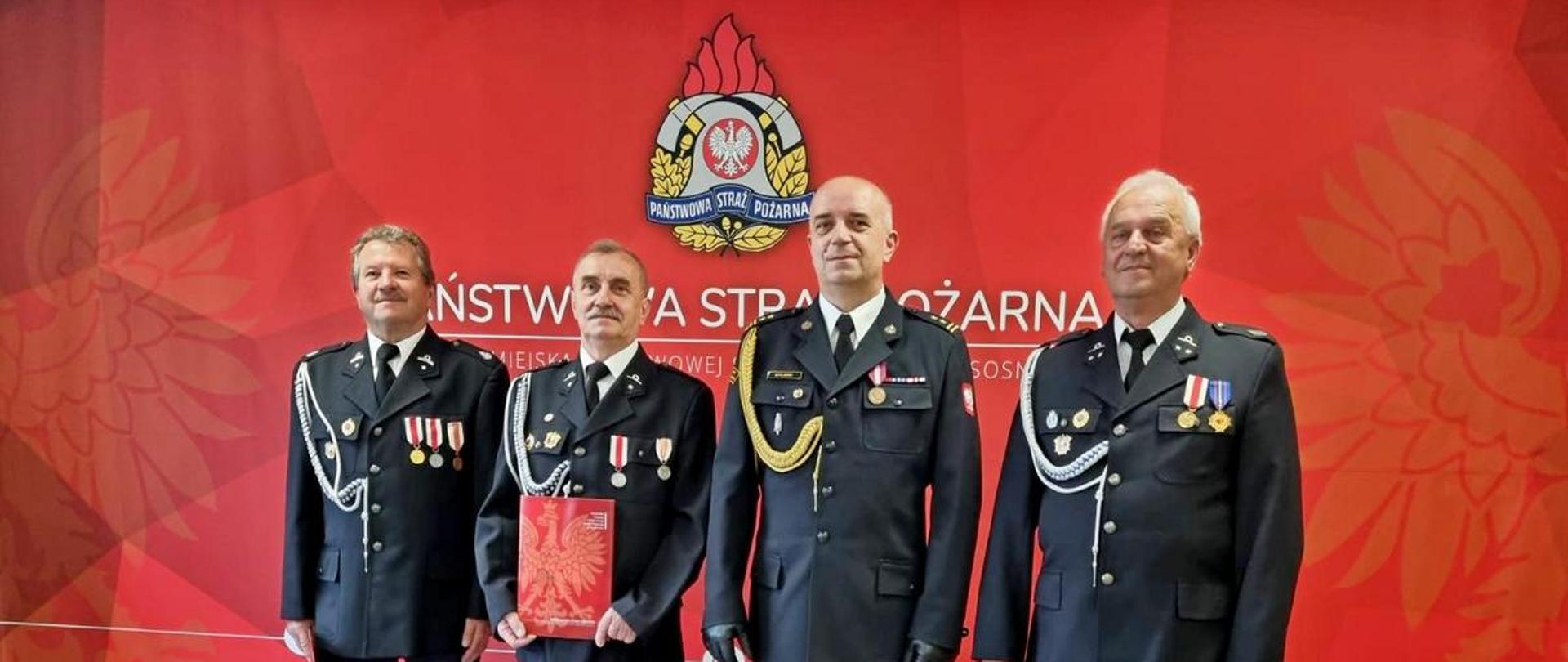Na zdjęciu Komendant Miejski Państwowej Straży Pożarnej w Sosnowcu w umundurowaniu galowym oraz strażacy OSP w umundurowaniu galowym. W tle widoczne logo Państwowej Straży Pożarnej na czerwonym tle.