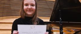 Dziewczynka stoi na estradzie sali koncertowej przed fortepianem i trzyma w rękach dyplom.