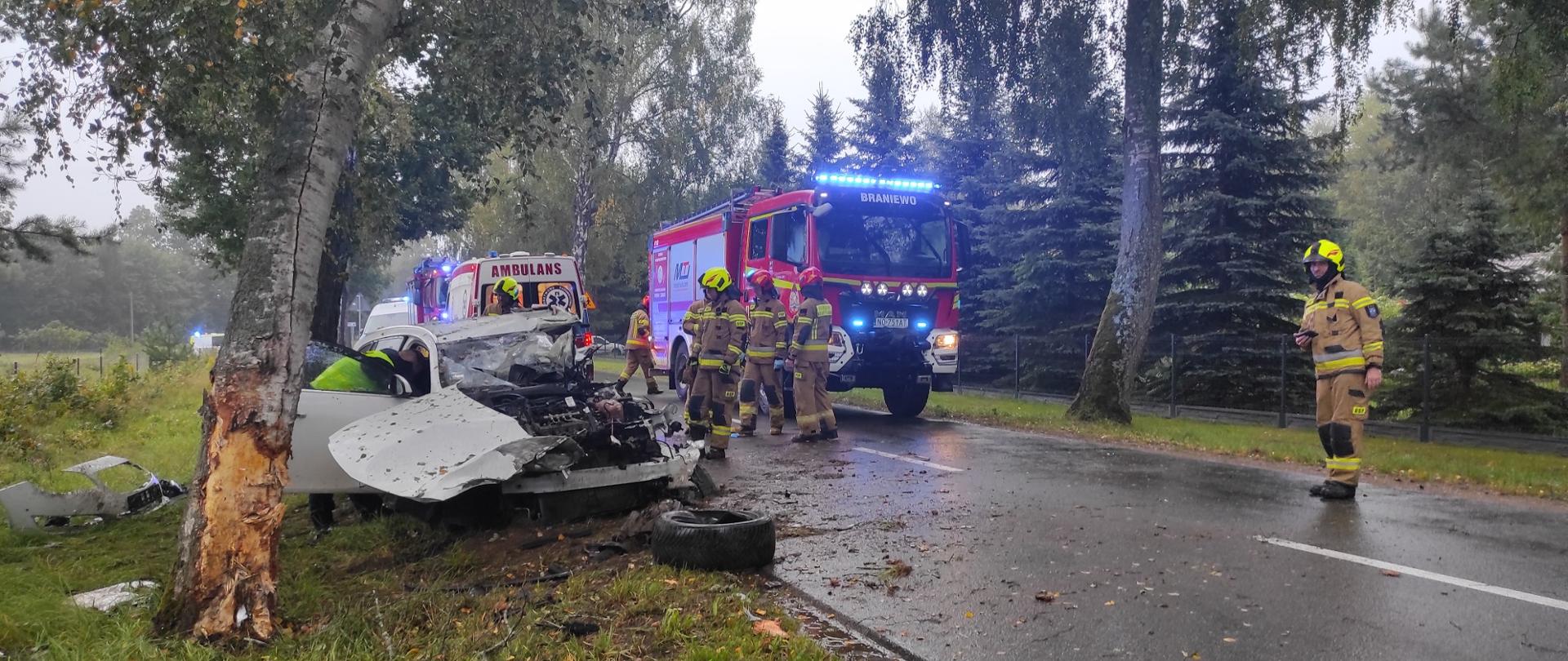 Zdjęcie przedstawia miejsce poważnego wypadku drogowego, który miał miejsce na wilgotnej, leśnej drodze. Na pierwszym planie widoczny jest mocno uszkodzony samochód osobowy, który uderzył czołowo w drzewo – uderzenie było bardzo silne, co widać po zniszczonym przodzie pojazdu i dużym złamaniu drzewa. Elementy karoserii oraz koło oderwane od auta leżą na poboczu.
Na miejscu zdarzenia pracują służby ratunkowe. Kilku strażaków w ubraniach ochronnych PSP znajduje się przy wozie strażackim. W tle widoczna jest karetka pogotowia z napisem a ratownicy udzielają pomocy osobie poszkodowanej – najprawdopodobniej kierowcy, którego strażak pochyla się wewnątrz rozbitego auta.
Cała akcja ratownicza odbywa się w deszczowych warunkach – jezdnia jest mokra, a otoczenie sugeruje, że do zdarzenia doszło na obrzeżach miejscowości. Zdjęcie pokazuje dynamiczne działanie służb i powagę sytuacji.
