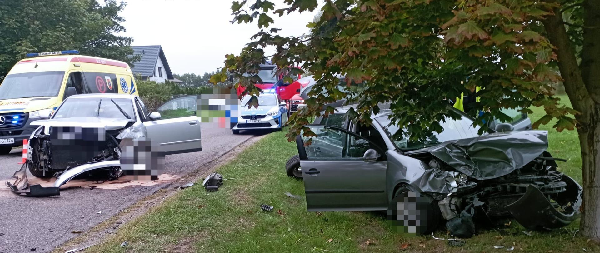 Na zdjęciu widać dwa rozbite samochody osobowe. Obydwa mają rozbite przody i otwarte drzwi. Jeden samochód stoi na poboczu obok drzewa a drugi na jezdni. W tle widać żółta karetkę pogotowia, radiowóz policyjny, dwa czerwone samochody straży pożarnej, dom oraz drzewa. Jest widno.