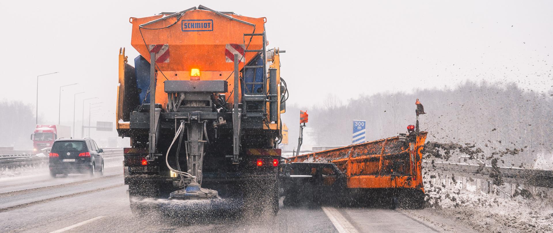 Pojazd do zimowego utrzymania dróg jedzie autostradą podczas opadów śniegu. Maszyna rozsypuje środek przeciwoblodzeniowy i odgarnia śnieg dużym bocznym pługiem, zrzucając mieszankę śniegu i błota na pobocze. W tle widoczne są samochody oraz zasypany krajobraz.