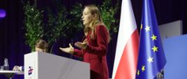 Minister funduszy i polityki regionalnej Katarzyna Pełczyńska-Nałęcz stoi na scenie za białą mównicą z napisem "EU Poland25.eu", za jej plecami flagi Polski i UE i zielone drzewka 