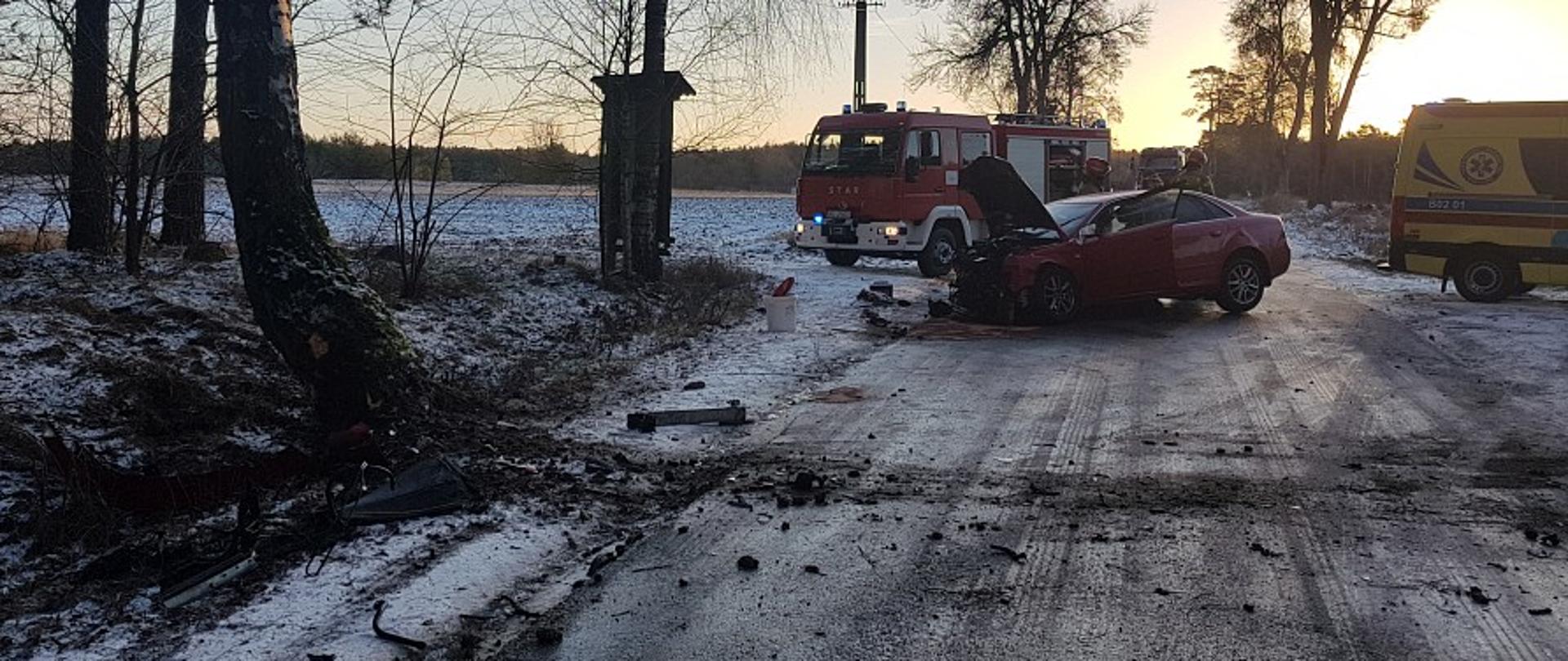 Na zdjęciu widać uszkodzony samochód osobowy stojący w poprzek jezdni, za nim znajdują się pojazdy służb ratowniczych.