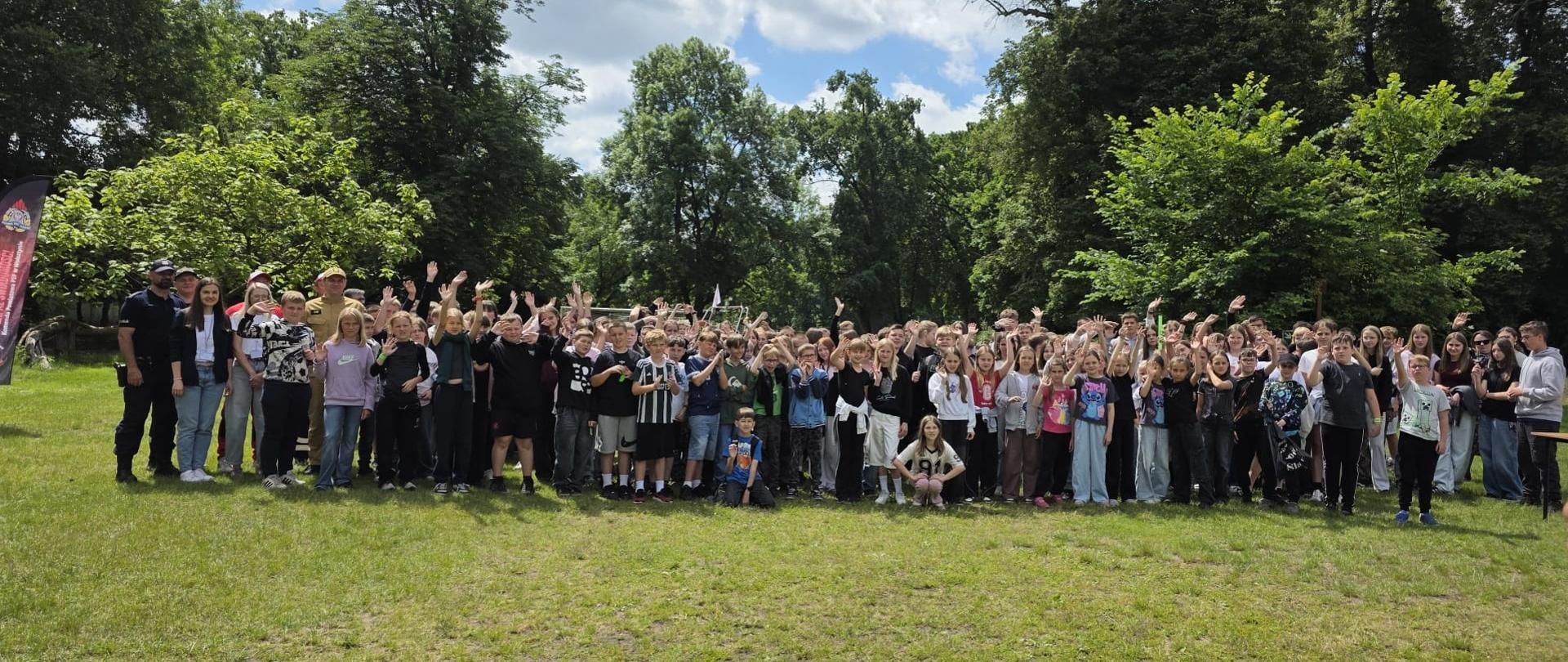 Zdjęcie grupowe dużej liczby dzieci, młodzieży i opiekunów na trawiastej polanie w otoczeniu wysokich drzew. Uczestnicy pozują do fotografii, część z nich macha do aparatu. W tle widoczne jest błękitne niebo z chmurami.