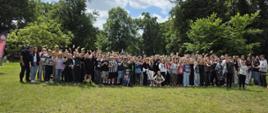 Zdjęcie grupowe dużej liczby dzieci, młodzieży i opiekunów na trawiastej polanie w otoczeniu wysokich drzew. Uczestnicy pozują do fotografii, część z nich macha do aparatu. W tle widoczne jest błękitne niebo z chmurami.