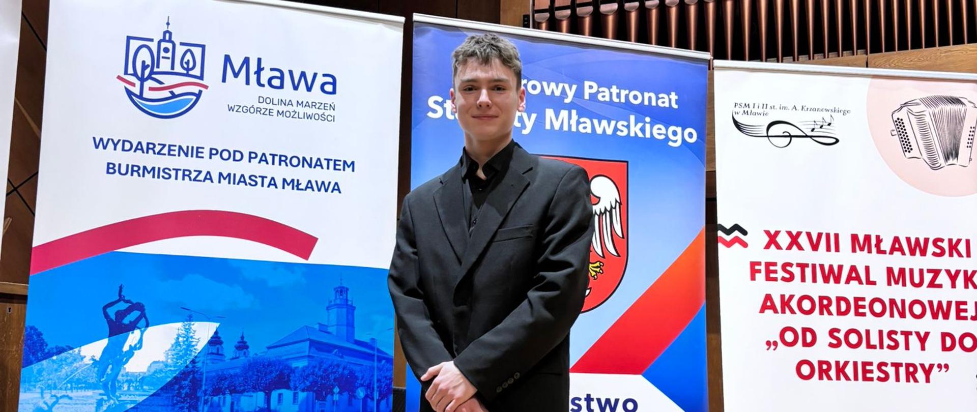 Zdjęcie - Mateusz Neckar w trakcie konkursu akordeonowego w Mławie. Uczeń na tle banerów reklamowych konkursu ubrany w czarny garnitur.