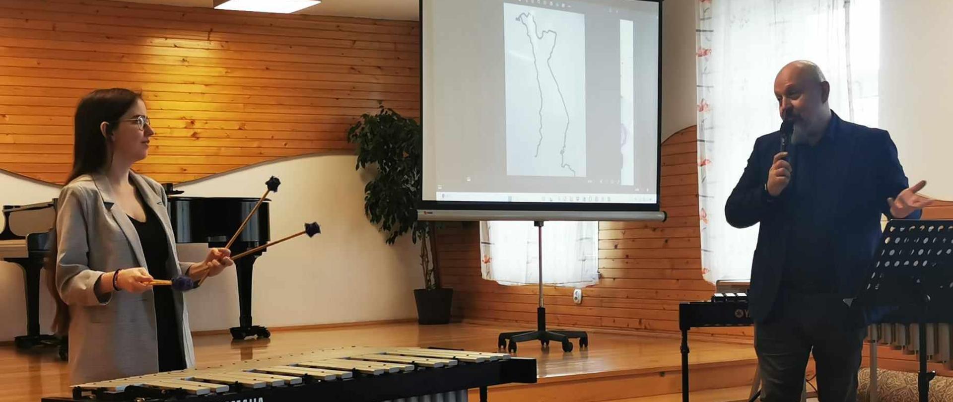 przed sceną nauczyciel zapowiadający prezentację oraz uczennica grająca na instrumencie perkusyjnym