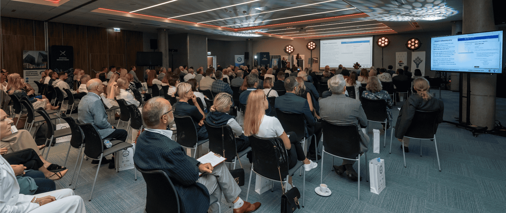 Fotografia przedstawia salę konferencyjną. Na pierwszym planie siedzą uczestnicy spotkania na krzesłach, odwróceni tyłem do zdjęcia. W oddali widać wyświetlaną prezentację na projektorze.