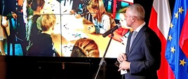 Minister cyfryzacji, Marek Zagórski, podczas konferencji prasowej przemawia do mikrofonu. W tle prezentacja multimedialna oraz flagi Polski i UE.