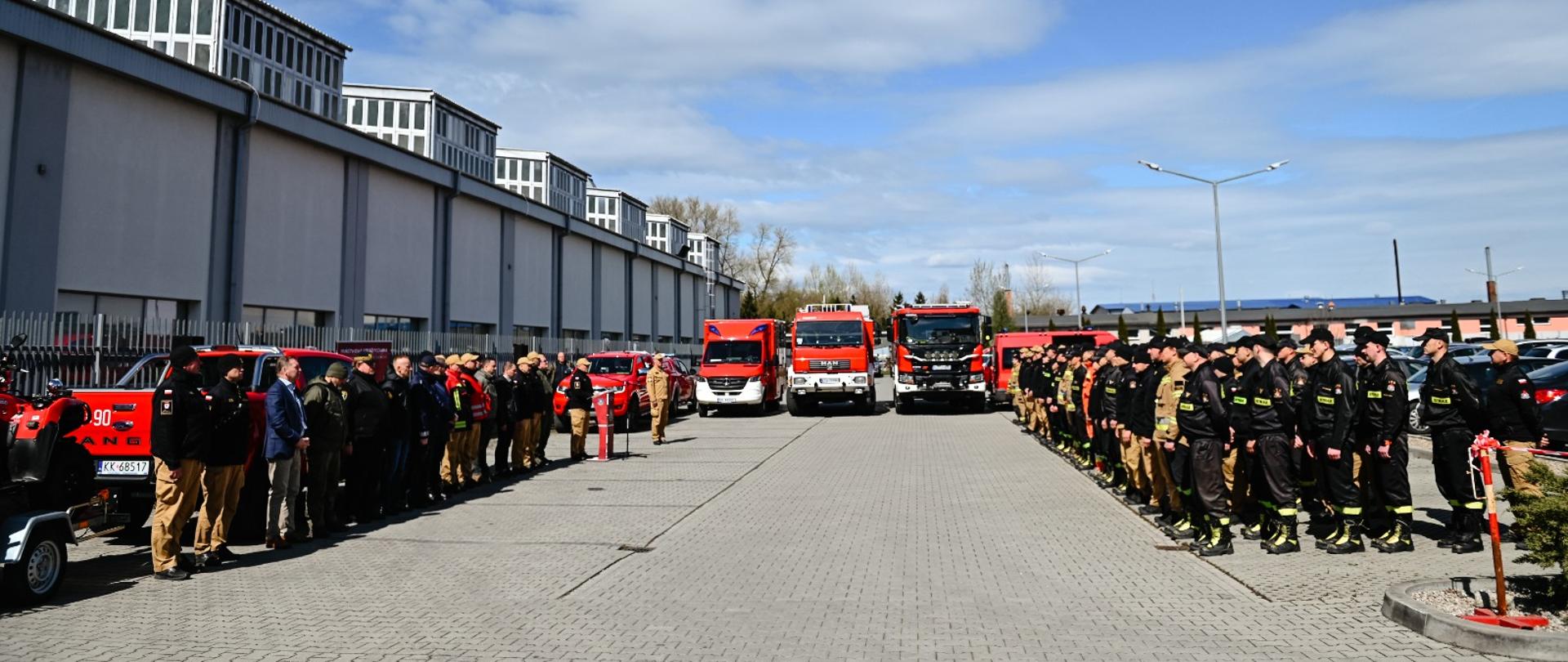 Strażacy oraz inni uczestnicy ćwiczeń stoją w szpalerach naprzeciw siebie na dużym parkingu wyłożonym szarą kostką. W tle samochody pożarnicze oraz budynki zakładów