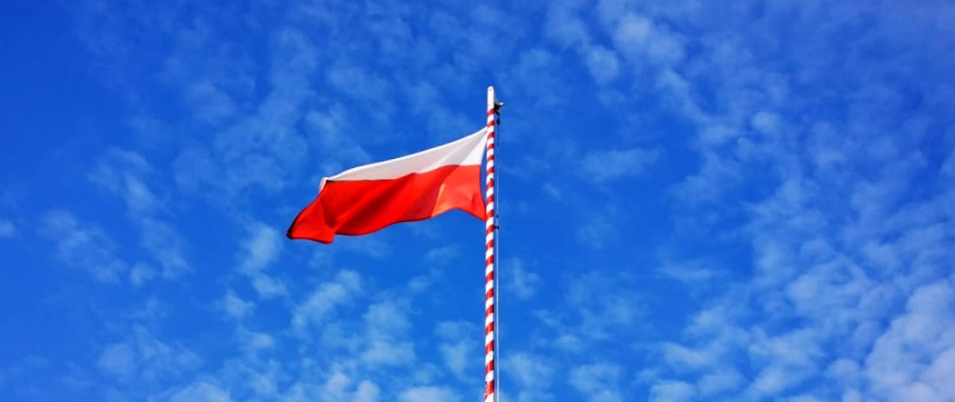 Zdjęcie przedstawia biało-czerwoną Flagę Rzeczypospolitej Polskiej łopoczącą na maszcie na tle błękitnego nieba.