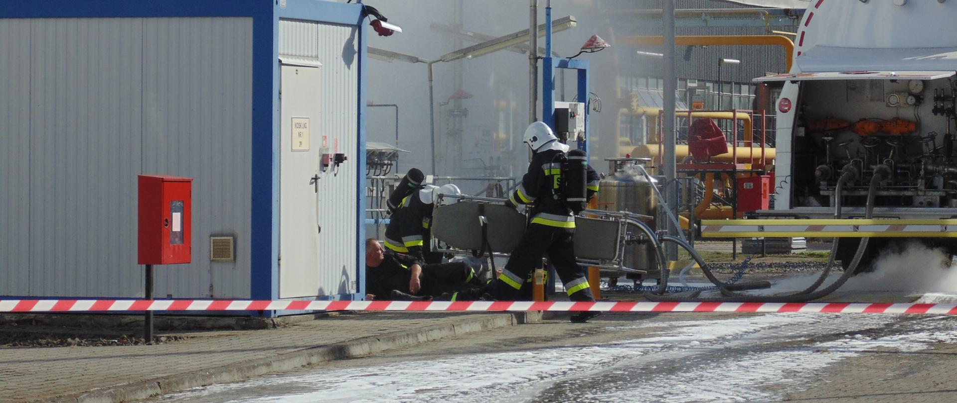 Wnętrze zakładu, strażacy udzielają pomocy poszkodowanemu mężczyźnie.