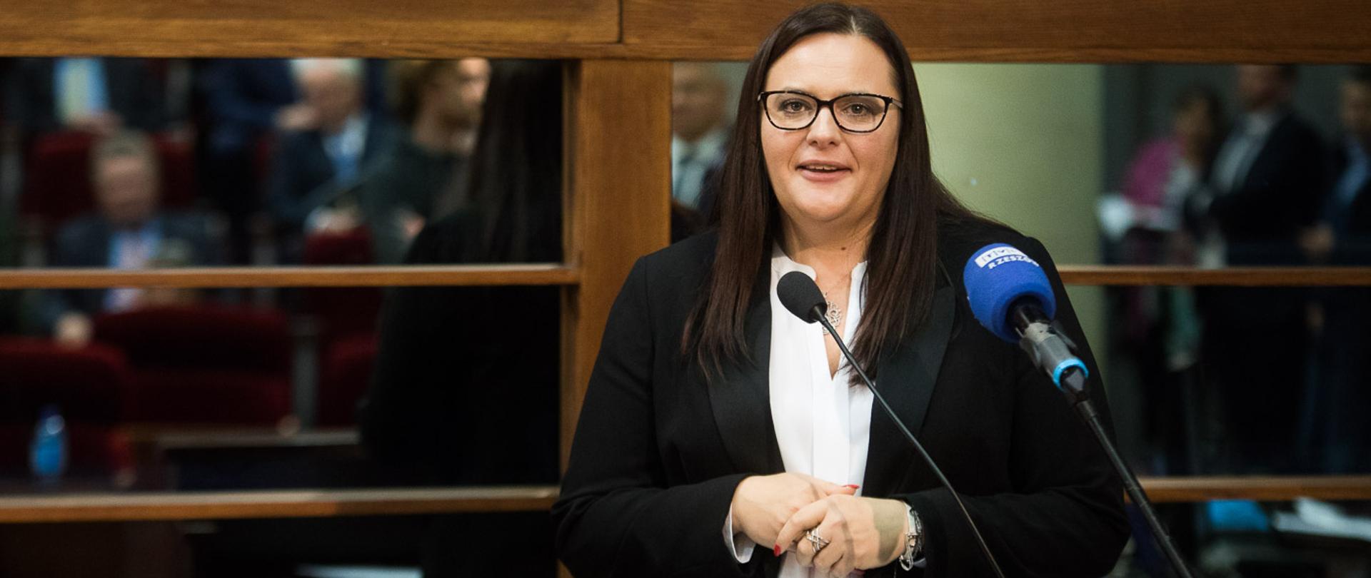 Uśmiechnięta minister Małgorzata Jarosińska-Jedynak stoi w mównicy i mówi do mikrofonów