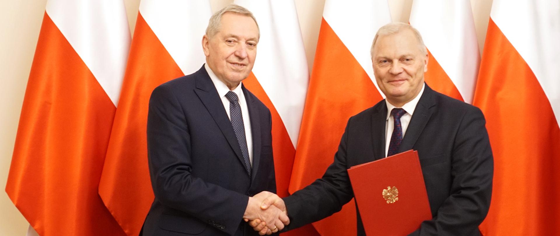 Wicepremier Henryk Kowalczyk i sekretarz stanu Lech Kołakowski podają sobie rękę na tle polskich flag.