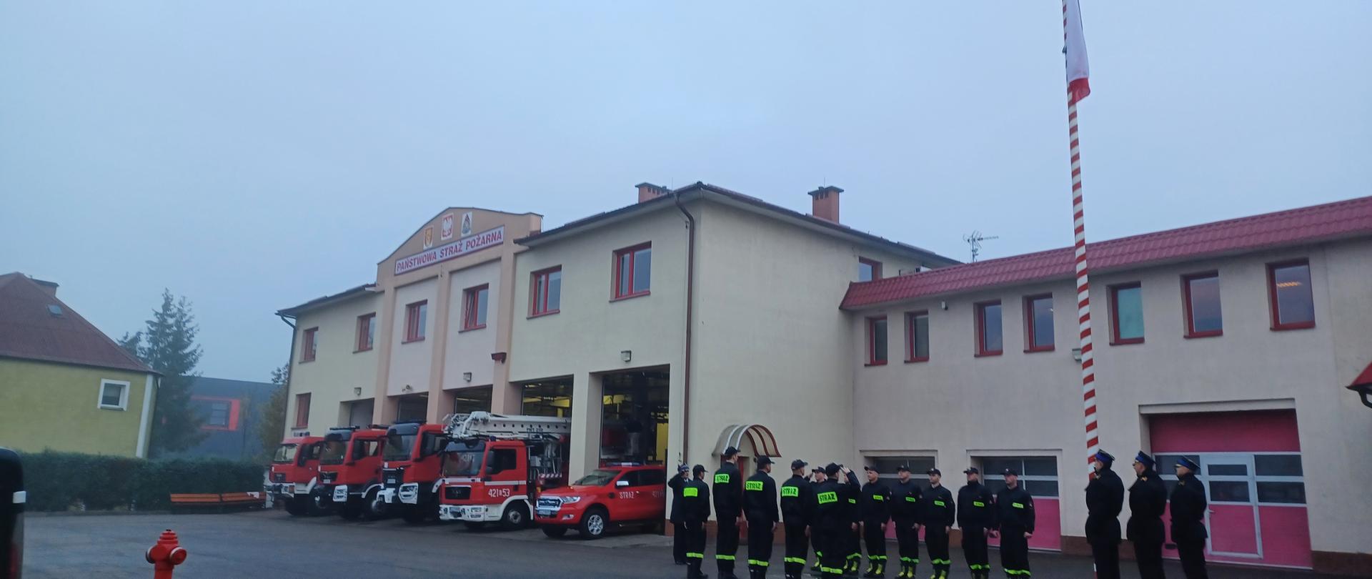 Uroczysta zmiana służby z okazji Narodowego Święta Niepodległości. Strażacy ze zmian służbowych stoją naprzeciwko siebie. Poczet flagowy wciąga flagę państwową na maszt. Komendant, poczet sztandarowy dowódcy zmian salutują. W tle budynek komendy i samochody pożarnicze.