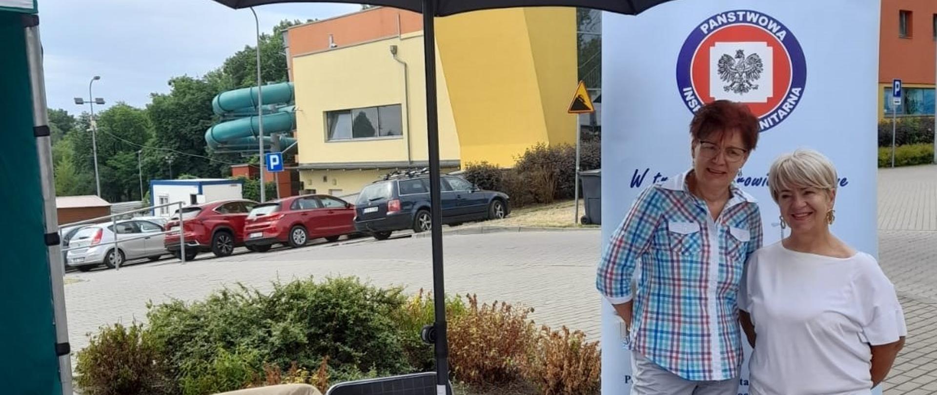 stoją dwie uśmiechnięte kobiety przy stoisku prezentującym działalność organizacji 