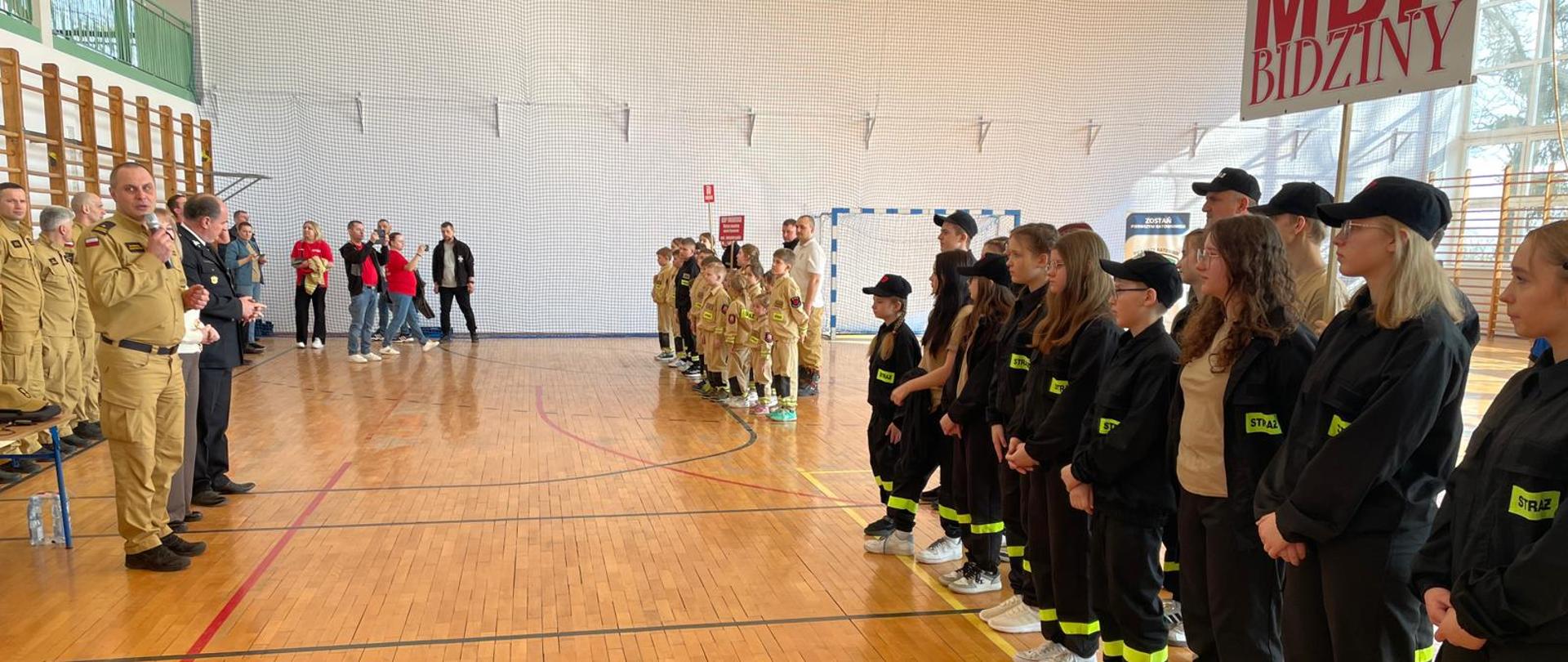 Na zdjęciu widoczne są po prawej stronie ustawione w szeregu cztery drużyny biorące udział w zawodach. Drużyny trzymają tabliczki z nazwami miejscowości, które reprezentują. Wysłuchują przemówienia funkcjonariuszy PSP. Oczekują na rozdanie nagród. Uczestnicy konkrecji ubrani są w czarne oraz żółte mundury. Funkcjonariusze PSP ubrani są w żółte mundury.