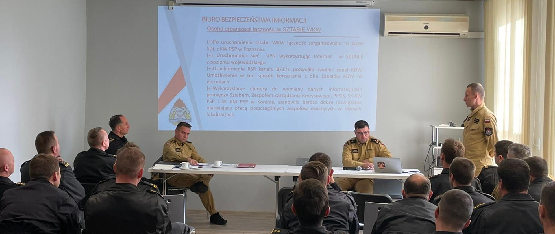 W oddali, na tle ekranu z wyświetlaną prezentacją, widoczny stolik przy którym dowództwo prowadzi przedmiotowe spotkanie. Omawiający prezentacje w pozycji stojącej. Widok na salę gdzie na krzesłach siedzi kilkunastu funkcjonariuszy. 