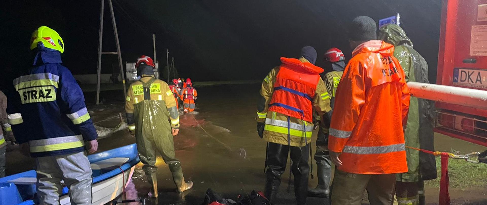 Grupa funkcjonariuszy straży pożarnej podczas działań na terenach dotkniętych sytuacją powodziową w tle widzimy wezbraną wodę. 