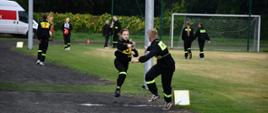 Zdjęcie przedstawia młodzieżową sztafetę strażacką rozgrywaną na bieżni stadionu podczas zawodów pożarniczych. W centrum kadru dziewczyna w czarnym mundurze strażackim z żółto-zielonymi odblaskami podaje prądownicę wodną jako pałeczkę sztafetową koledze z drużyny. Na ich plecach widoczne są żółte plastrony z numerami startowymi. Oboje biegaczy uchwyconych jest w dynamicznym ruchu na żwirowej bieżni, oddzielonej krawężnikiem od trawiastego boiska. W tle widać bramkę piłkarską, siatkę ochronną oraz innych uczestników zawodów i osoby towarzyszące, z których część również nosi strażackie umundurowanie lub ubrania z numerami. Z lewej strony stoi zaparkowany biały samochód dostawczy. Całe wydarzenie odbywa się w otoczeniu zieleni drzew.