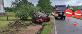 Zdjęcie wykonane w dzień. W tle pochmurne niebo . Na poboczu drogi , na trawniku łączącym się z drogą leży na dachu małe auto osobowe. Jest zniszczone z każdej strony.