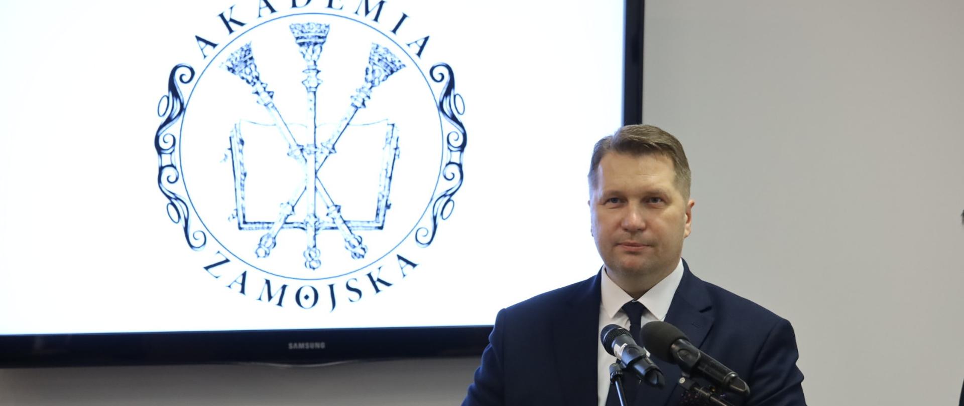 Uroczysta inauguracja roku akademickiego na Akademii Zamojskiej z udziałem ministra Przemysława Czarnka. minister przemawia do mikrofonu przy mówinicy. w tle logo akademii zamojskiej