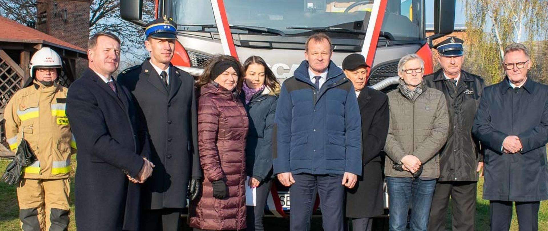 zdjecie grupowe strażaków, kobiet, mężczyzn na tle samochodu strażackiego, ktory na przedniej szybie ma bialło czerwoną wstegę, w tle murowane budowle