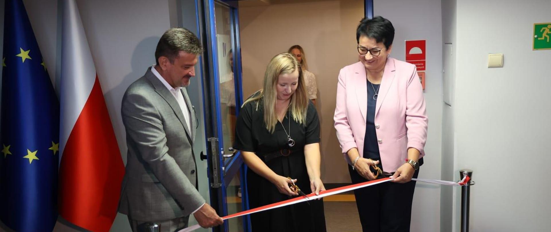 Wojewoda Opolski Monika Jurek, Przewodnicząca Wojewódzkiego Zespołu ds. Orzekania o Niepełnosprawności Magdalena Kowalewska oraz Wicewojewoda Piotr Pośpiech przecinają wstęgę przed wejściem do nowej siedziby WZON. Po lewej stronie widać flagi Polski oraz Unii Europejskiej.