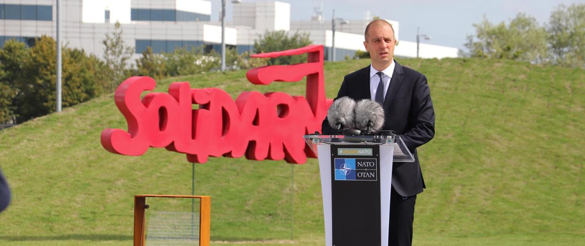 Ambasador Tomasz Szatkowski przemawia na tle logo Solidarności przed Kwaterą Główną NATO