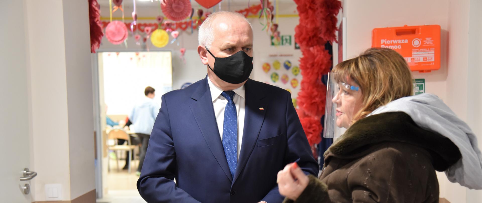 Wojewoda z panią wójt stoją w jasnym, dobrze oświetlonym korytarzu. Za nimi znajduje się ustrojona sala. Daleko w tle widać stojącą tyłem kobietę 