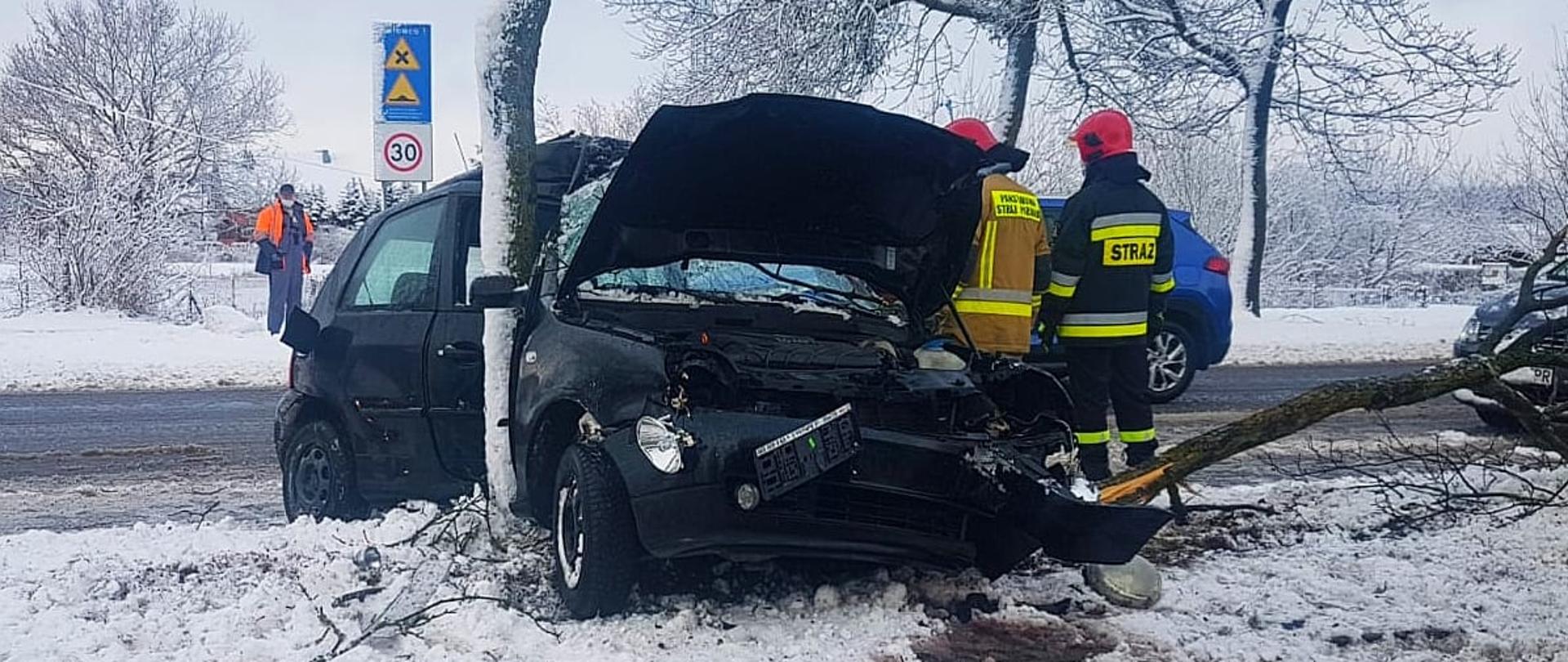Zdjęcie przedstawia samochód osobowy, który uderzył w drzewo, uszkodzony bok auta, kolor auta czarny.
