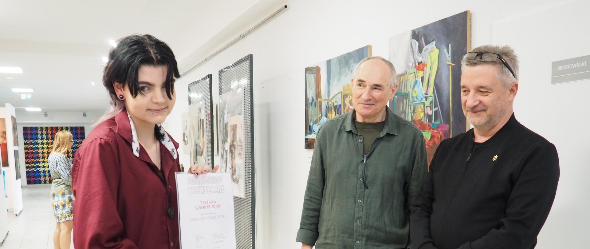 Na zdjęciu widzimy młodą osobę w bordowej koszuli, trzymającą w rękach dyplom z napisem „Tatiana Grobelna”. Stoi ona we wnętrzu galerii lub szkoły artystycznej, w otoczeniu dwóch uśmiechniętych mężczyzn – prawdopodobnie nauczycieli lub jurorów. W tle znajdują się obrazy zawieszone na ścianie oraz inne osoby oglądające wystawę. Całość sprawia wrażenie uroczystości wręczenia nagród lub zakończenia programu artystycznego. Atmosfera jest przyjazna i pełna dumy. Od lewej: Tatiana Grobelna, Zdzisław Kwiatkowski, dyr. Krzysztof Dąbek.

