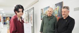 Na zdjęciu widzimy młodą osobę w bordowej koszuli, trzymającą w rękach dyplom z napisem „Tatiana Grobelna”. Stoi ona we wnętrzu galerii lub szkoły artystycznej, w otoczeniu dwóch uśmiechniętych mężczyzn – prawdopodobnie nauczycieli lub jurorów. W tle znajdują się obrazy zawieszone na ścianie oraz inne osoby oglądające wystawę. Całość sprawia wrażenie uroczystości wręczenia nagród lub zakończenia programu artystycznego. Atmosfera jest przyjazna i pełna dumy. Od lewej: Tatiana Grobelna, Zdzisław Kwiatkowski, dyr. Krzysztof Dąbek.
