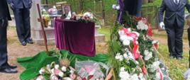 Ceremonia pogrzebowa na cmentarzu. Posterunek honorowy strażaków stoi obok urny przed złożeniem jej w miejsce ostatniego spoczynku. Obok urny stoją wieńce kwiatów oraz zdjęcie zmarłego.