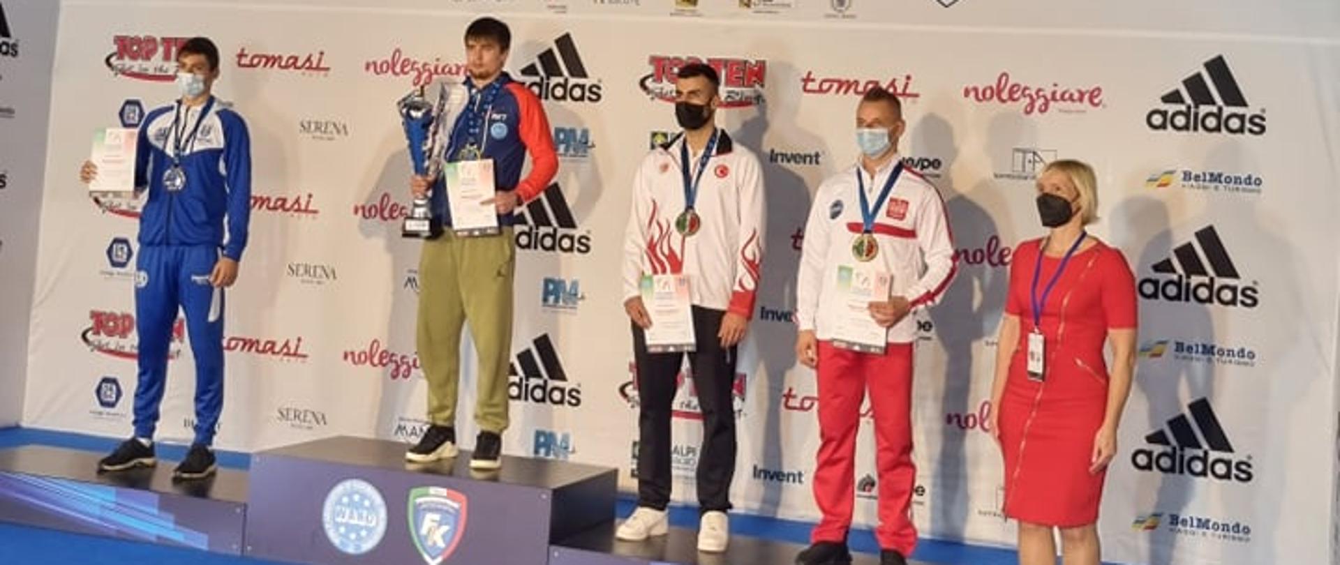 Na niebieskiej scenie znajduje się podium, a na nim zawodnicy z medalami na szyi. Adrian Durma stoi po prawej stronie ze srebrnym medalem i pamiątkowym dyplomem. Za plecami zawodnicy mają baner reklamowy.