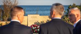 The photo shows three men standing with their backs to the camera (Minister Tomasz Siemoniak is in the center). The men are standing in front of the gravestone of Polish engineer Paweł Potocki. The gravestone consists of three plaques in different languages. Beneath the central plaque, inscribed in Polish, lies a wreath in red (roses), white (gerberas), and green, with a white-and-red sash. To the right and left of the gravestone plaques, vegetation is visible, with the blue sea in the background. 