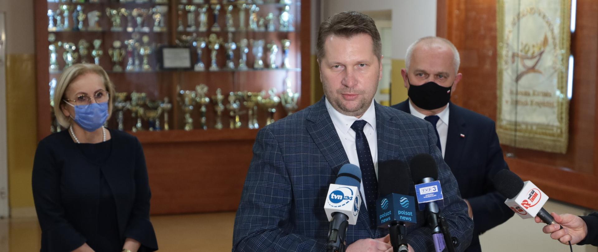 minister udziela wywiadu do mikrofonu, za nim inni uczestnicy spotkania i gablota z pucharami