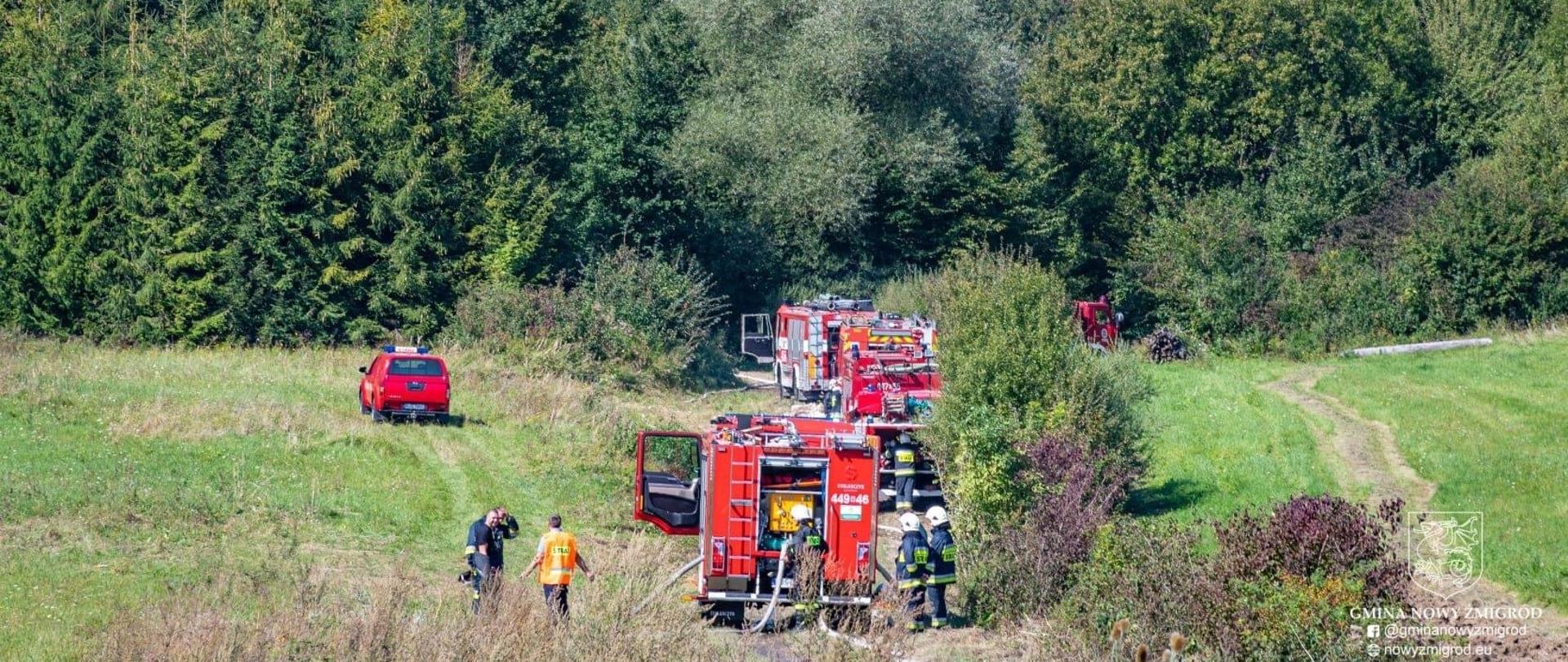 Na zdjęciu samochody ratowniczo-gaśnicze pod lasem na łące przygotowujące się do przyjęcia wody z dwóch magistrali wodnych. Dookoła samochodów stoją strażacy z OSP z terenu Gminy Nowy Źmigród