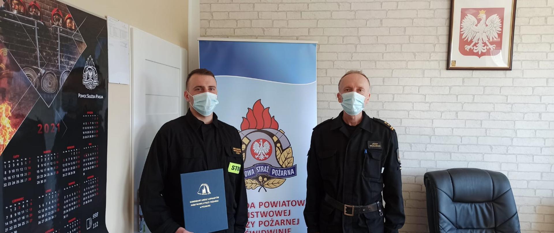 Zdjęcie przedstawia dwóch mężczyzn ubranych w czarne mundury strażackie oraz maseczki chirurgiczne, jeden z nich trzyma w rękach dokumenty. Za nimi widnieje baner z logiem PSP oraz napisem: "Komenda Powiatowa Państwowej Straży Pożarnej w Świdwinie". Za banerem widzimy ścianę z tapetą w szare cegły. Na ścianie wisi Godło Rzeczypospolitej Polskiej. Zdjęcie wykonano w gabinecie Komendanta Powiatowego PSP w Świdwinie.