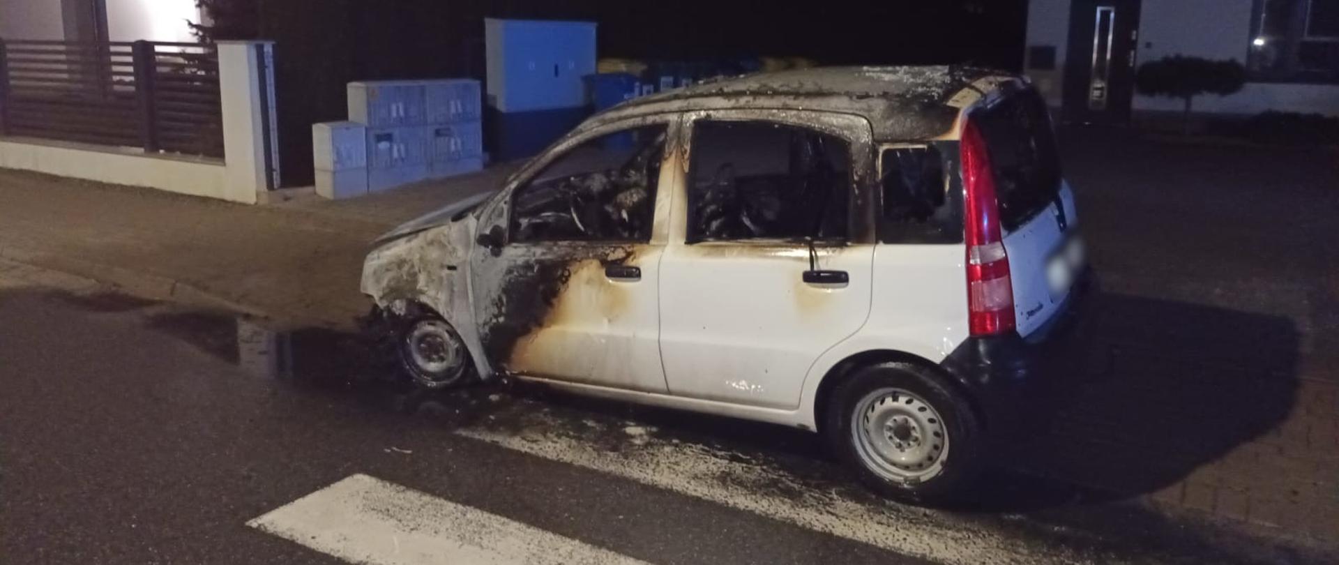 Widoczne miejsce zdarzenia oraz wrak samochodu marki FIAT Panda w kolorze białym po pożarze.