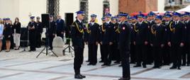 D-ca uroczystości ubrany w mundur galowy składa meldunek Komendantowi Wojewódzkiemu. Z tyłu ustawienie w kilku szeregach odznaczeni strażacy również ubrani w mundury galowe w tle ratusz