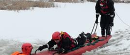 Zdjęcie przedstawia strażaków podczas działań ratowniczych na zamarzniętym zbiorniku wodnym. Po lewej stronie zdjęcia strażak w przeręblu wodnym w pomarańczowym ubraniu wodnym z czerwonym kaskiem. Po prawej stronie dwóch strażaków w czarnych kombinezonach wodnych z czerwonymi hełmami zbliżają się do przerębla na pomarańczowych saniach lodowych. Jeden ze strażaków leży na brzuchu z lewej strony sań twarzą do przerębla, drugi w pozycji stojącej popycha sanie w kierunku przerębla. Tafla lodu na akwenie jest pokryta śniegiem