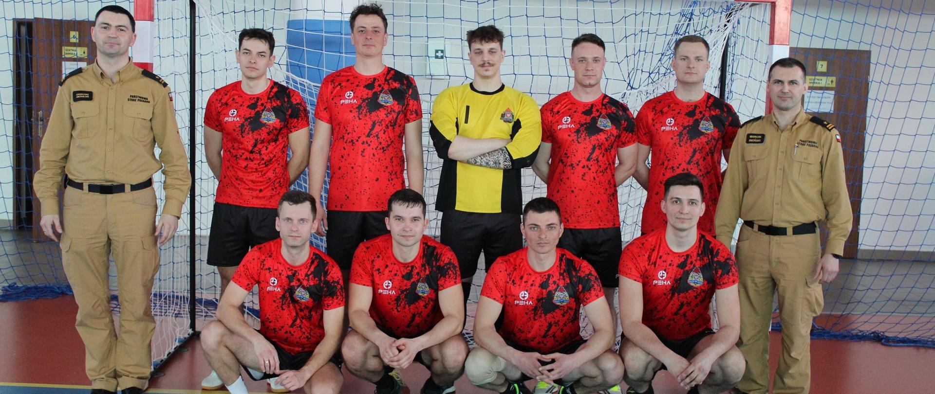 Na zdjęciu widać dziesięciu mężczyzn ustawionych do wspólnego zdjęcia w hali sportowej.
Czterech z nich klęczy z przodu, a siedmiu stoi z tyłu. Większość ma na sobie czerwone koszulki sportowe z herbem straży pożarnej i czarne spodenki. Jeden z mężczyzn stojących z tyłu ma żółtą koszulkę bramkarską, dwóch po prawej i po lewej stronie beżowy mundur strażaka.