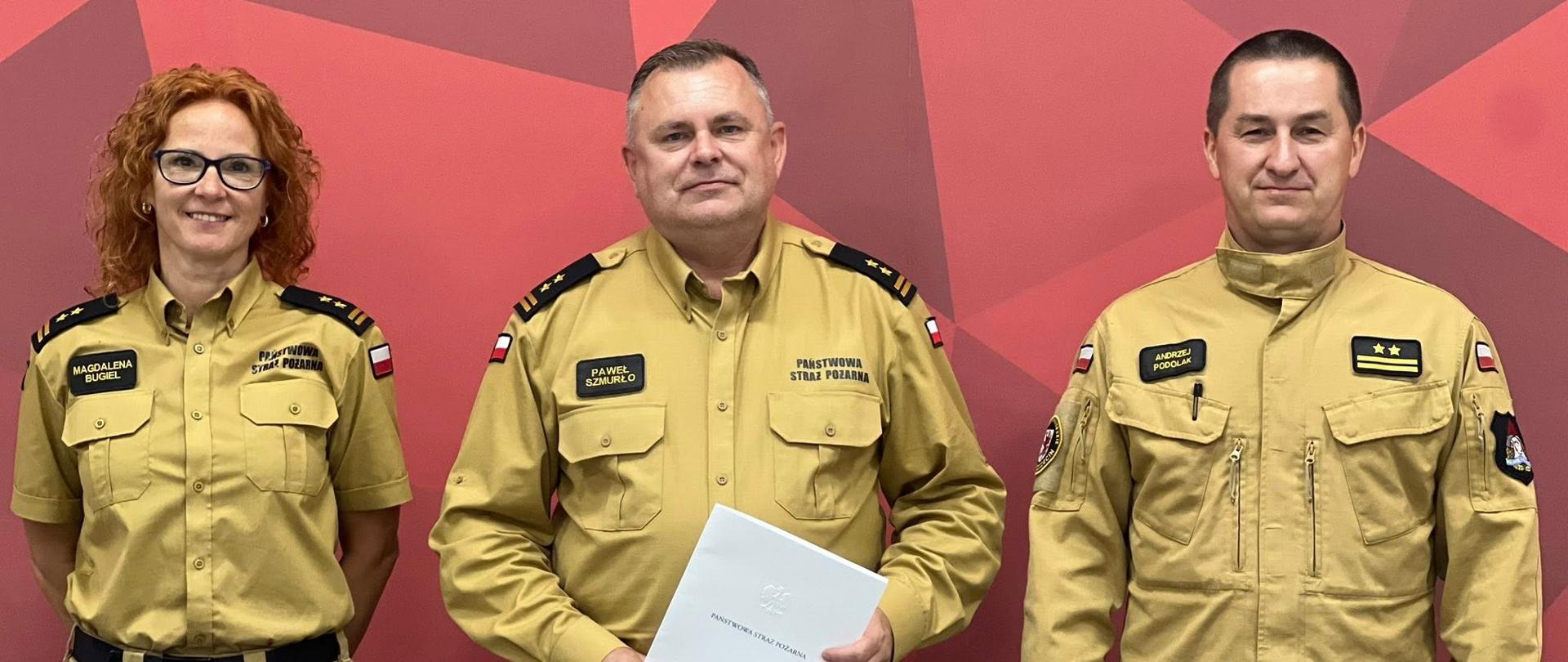 Trzy osoby stoją obok siebie na tle bordowej ściany. Ubrane są piaskowe mundury. Po lewej stronie stoi kobieta, która ma na lewej piersi naszywkę z imieniem i nazwiskiem. Kobieta ma rude, kręcone włosy i nosi okulary. Pośrodku stoi mężczyzna trzymający w rękach białą, złożoną kartkę. Na jego piersi widnieje naszywka z imieniem i nazwiskiem. Poniżej naszywki jest napis PAŃSTWOWA STRAŻ POŻARNA. Po prawej stronie stoi mężczyzna. Wszystkie trzy osoby noszą mundury z emblematami na ramionach i klapach, a także paski na wysokości pasa