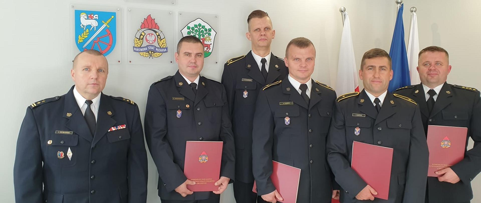 Na zdjęciu sześciu strażaków w gabinecie komendanta w mundurach wyjściowych. W rękach trzymają czerwone teczki.