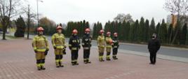 Poddodział strażaków podczas obchody upamiętnienia 102. Rocznicy Odzyskania Niepodległości. Po lewej stronie stoją strażacy w ubraniu specjalnym z zasłoniętymi twarzami i nosem. Po prawej stronie Dowódca Jednostki Ratowniczo-Gaśniczej w umundurowaniu wyjściowym w maseczce.