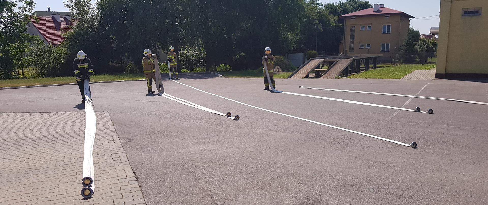 Zdjęcie przedstawia 4 strażaków zwijających węże pożarnicze. W tle widać plac wewnętrzny Komendy Powiatowej PSP w Sandomierzu