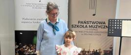 Zdjecie przedstawia uczennice w białej sukience oraz nauczycielke ubrana w czarne spodnie, błekitny sweterek na szyi przywiazana niebieska chustka. W tle widac plakat z napisem Państwowa Szkoła Muzyczna w Zielonej Górze