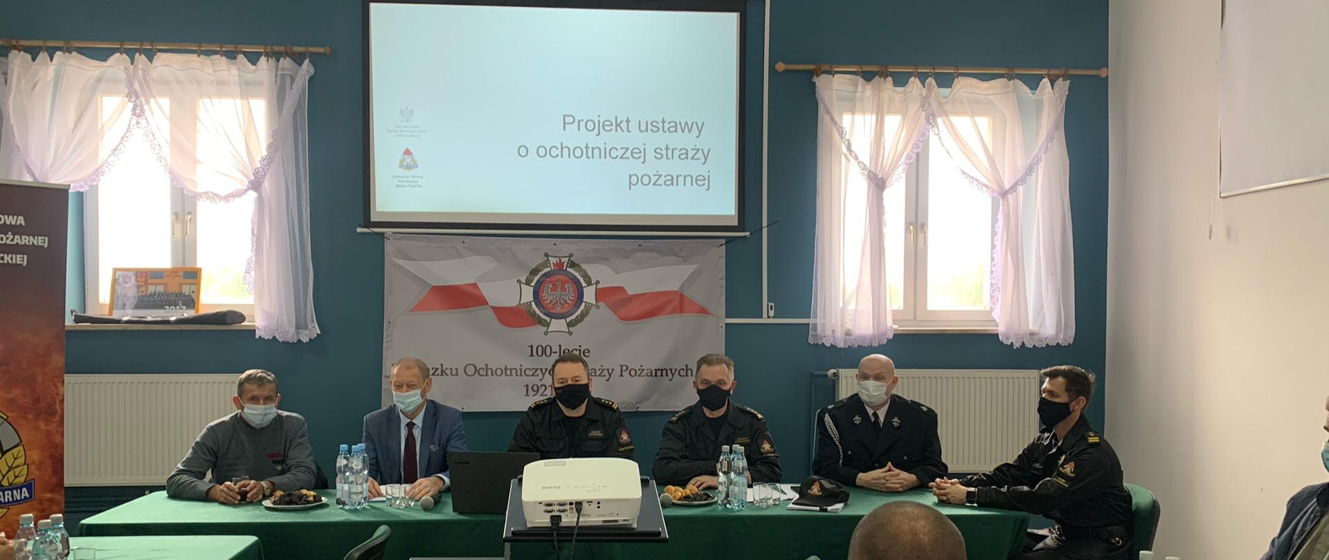 Zdjęcie przedstawia 6 mężczyzn siedzących za stołem, nakrytym zielonym obrusem. Za nimi znajduje się niebiska ściana z dwoma oknami, a pomiędzy nimi jest rzutnik, na którym wyświetlana jest prezentacja. Czterej mężczyźni od prawej strony ubrani są w staje dowódczo sztabowe, reszta mężczyzn ubrana jest po cywilu. Na stole znajdują się butelki z wodą oraz szklani i ciastka.